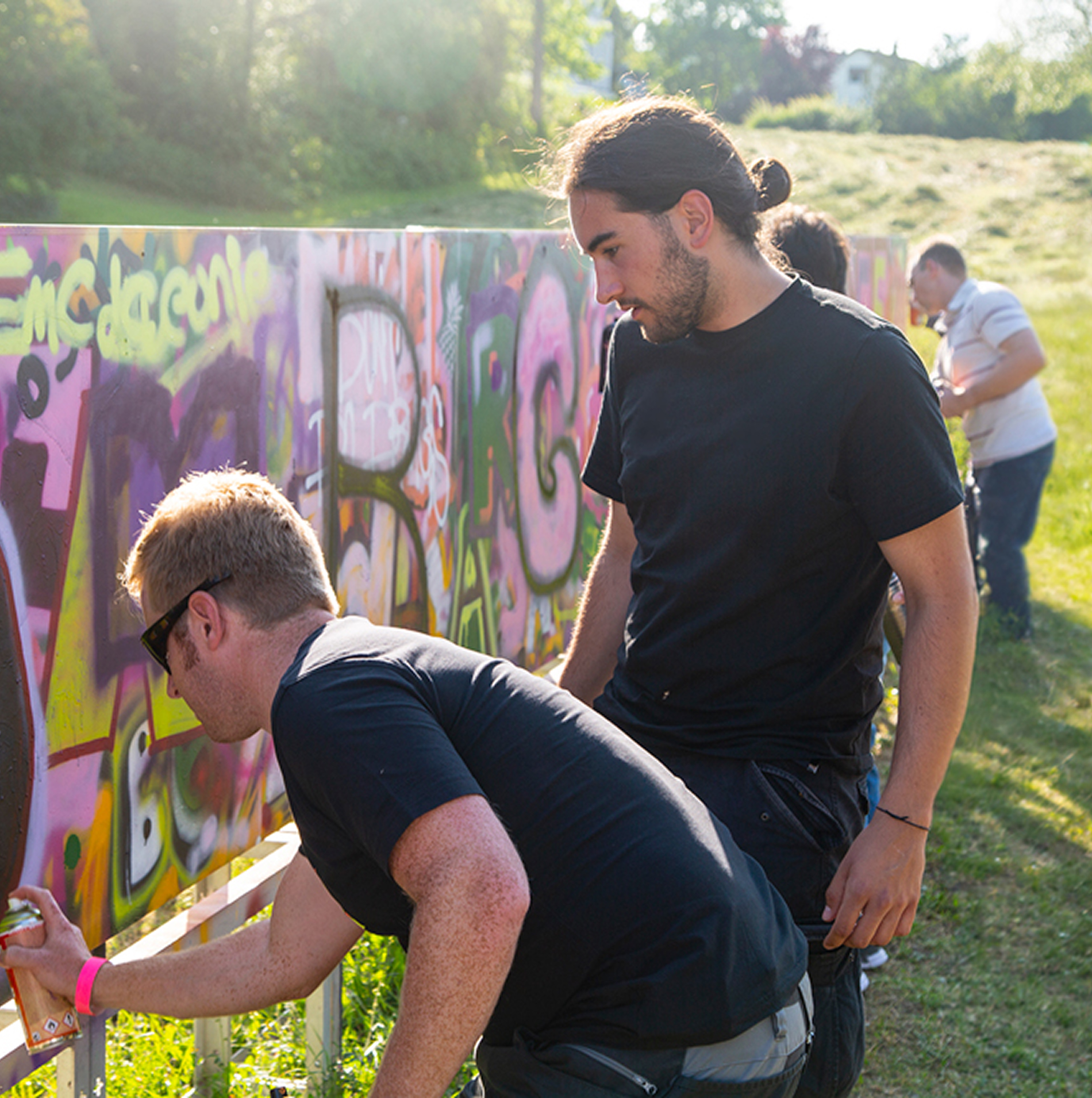 Personen gestalten gemeinsam eine bunte Graffiti-Wand im Freien auf einer Grünfläche. Der Moderator Colin Jäck erklärt einem Teilnehmer etwas.