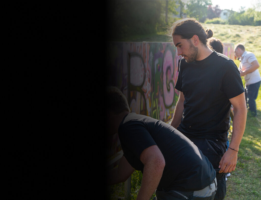 Personen gestalten gemeinsam eine bunte Graffiti-Wand im Freien auf einer Grünfläche. Der Moderator Colin Jäck erklärt einem Teilnehmer etwas.