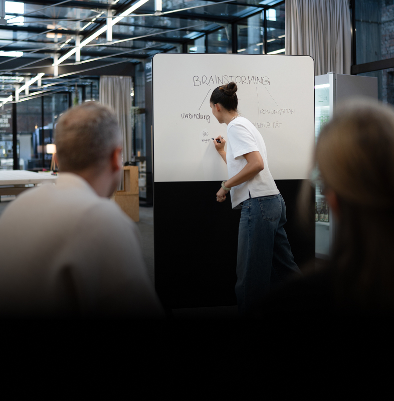Frau schreibt Begriffe als Brainstorming auf ein Whiteboard vor Zuhörenden.