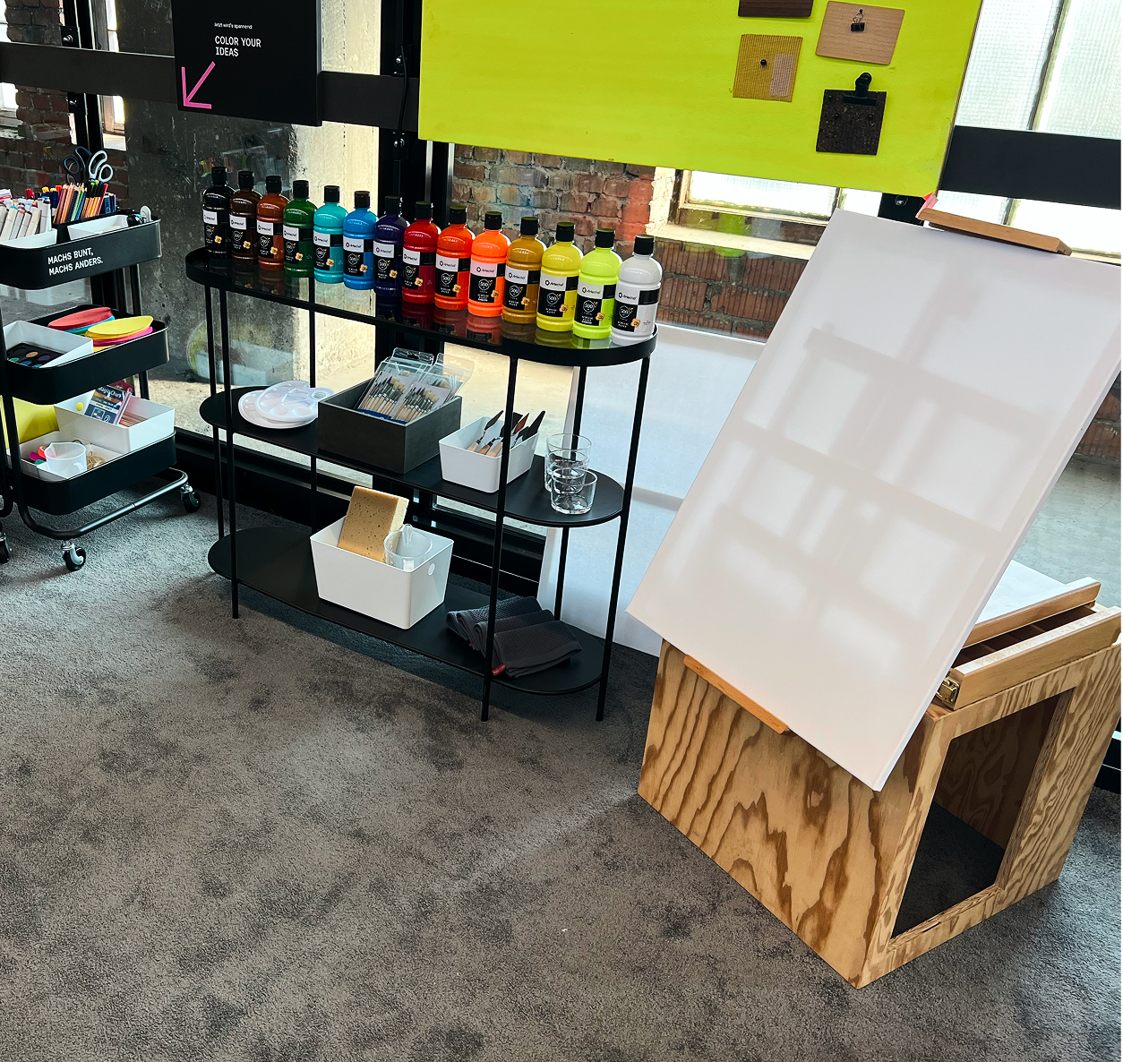 Malbereich mit Regal voller Farbflaschen, Pinseln und Utensilien neben einer leeren Leinwand auf einer Holzstaffelei, Schild „Color your ideas“