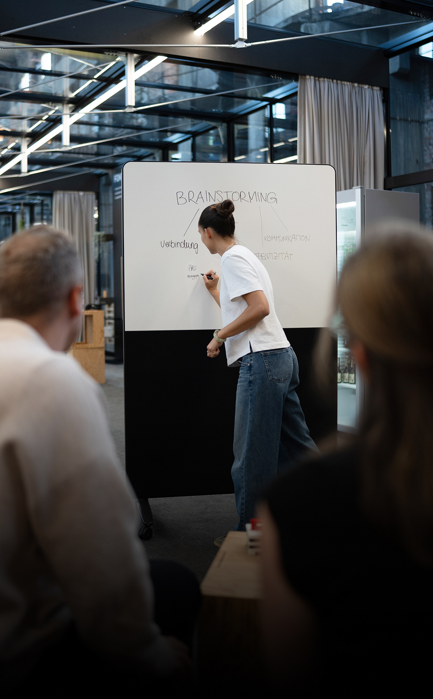 Frau schreibt Begriffe als Brainstorming auf ein Whiteboard vor Zuhörenden.