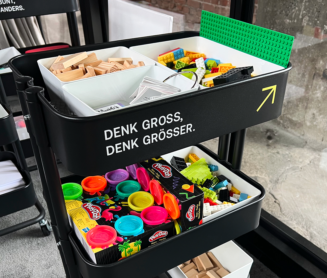 Rollwagen mit Kreativmaterial wie Bauklötzen, Lego und Play-Doh, beschriftet mit „Denk gross, denk grösser.“