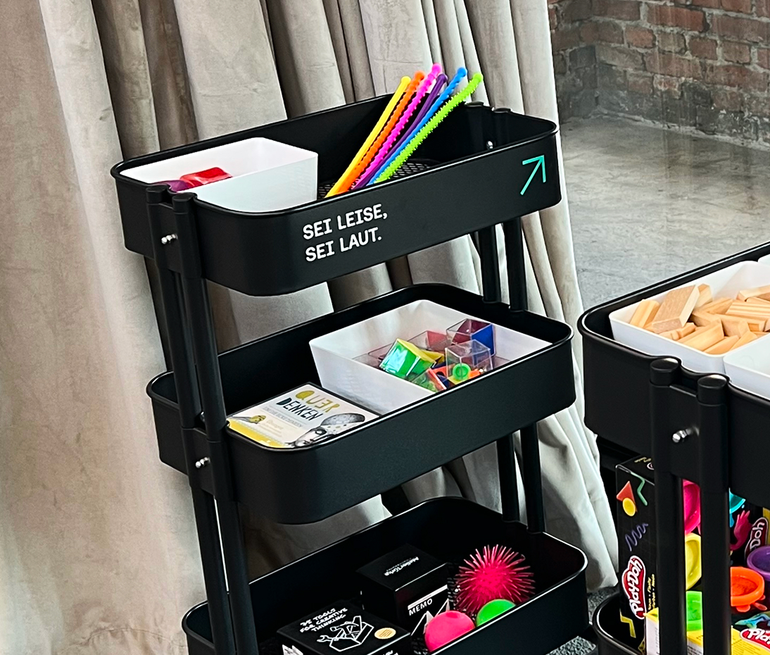 Rollwagen mit Aufschrift „Sei leise, sei laut“, gefüllt mit Sensorik-Spielzeug, Denkspielen und Knetmasse.