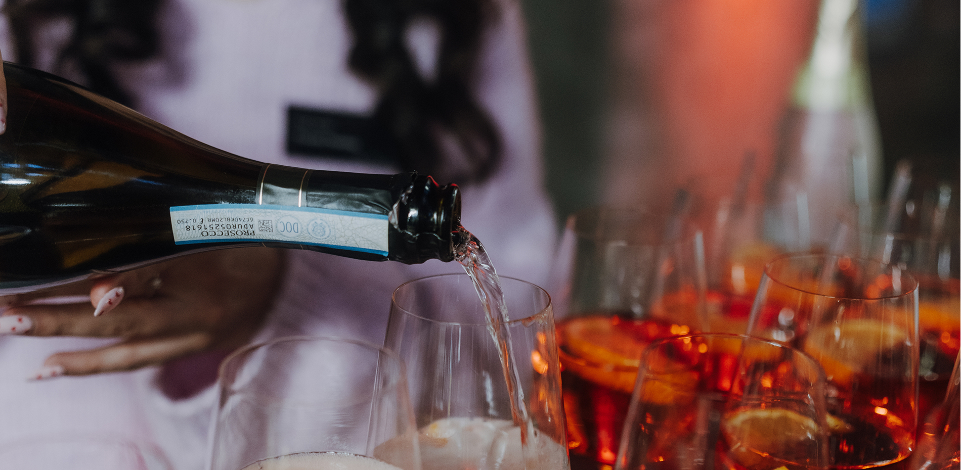Prosecco wird aus einer Flasche in ein Glas gegossen, umgeben von weiteren Gläsern mit Aperol Spritz